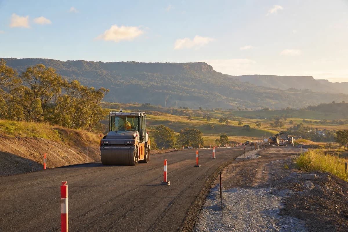 Roadworks Illawarra access road with roller compacting pavement against green foothills at late afternoon