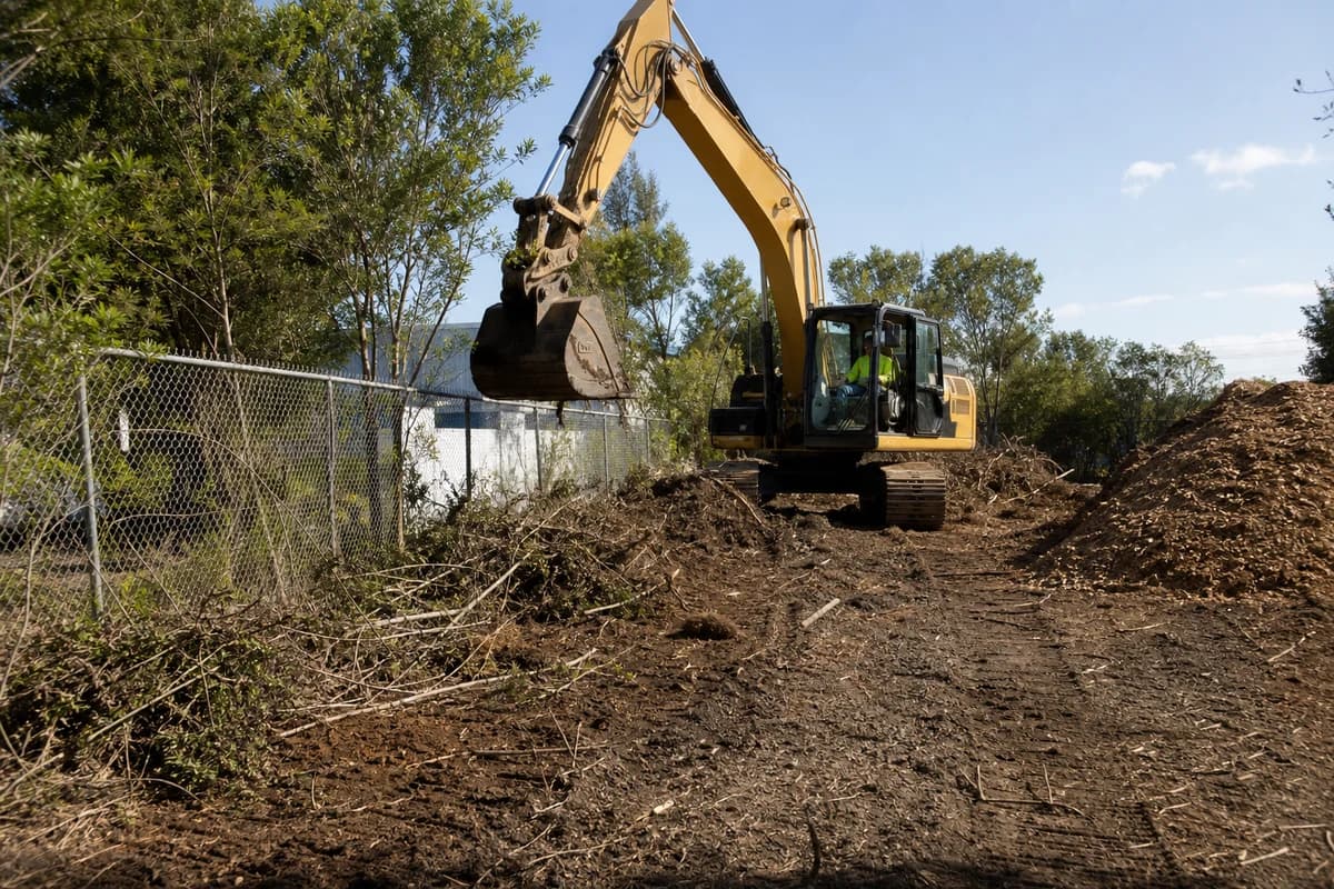 Site development civil works NSW clearing strip with excavator removing vegetation beside a fenced industrial lot at mid-morning
