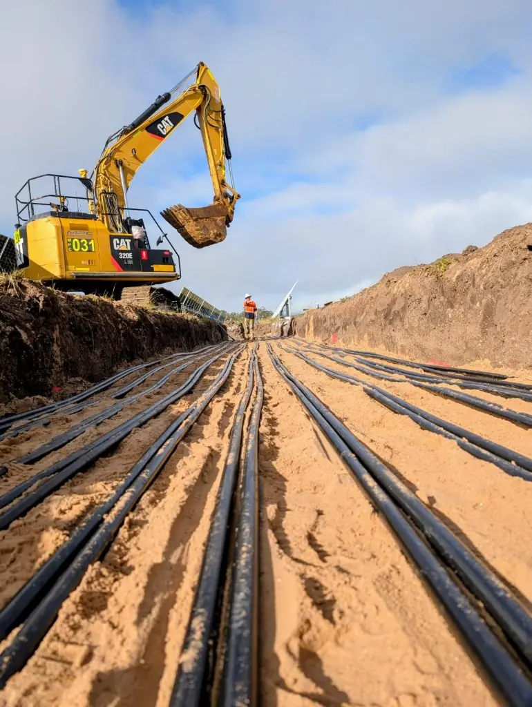 Excavator digging a renewable energy cable trench with conduits staged nearby.
